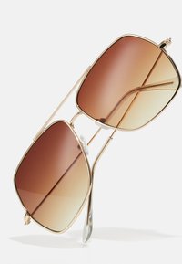 Gafas de sol cuadradas con un marco de metal dorado, lentes marrón degradado y almohadillas nasales de silicona transparentes. Patillas elegantes con un diseño minimalista.