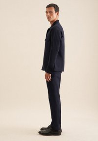 Marineblauwe, knoopsluitingjas met een borstzak, gecombineerd met bijpassende broek en zwarte schoenen, tegen een neutrale achtergrond.