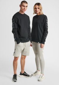 Sudadera negra con estampado de flechas en las mangas, combinada con pantalones cortos grises. Mujer en leggings grises. Ambas llevan zapatillas de colores coordinados.