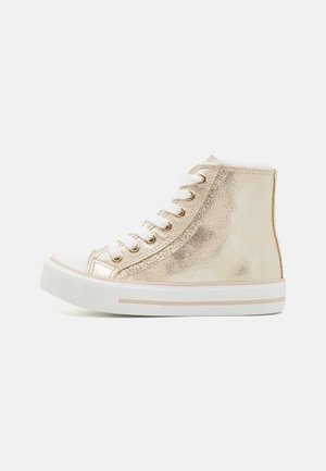 Basket montante avec dessus métallique doré brillant, lacets blancs, œillets dorés, et semelle en caoutchouc blanche avec bande beige sur fond clair.