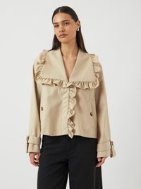 Femme portant une veste beige avec un grand col à volants et devant plissé, associée à un pantalon noir, debout devant un fond clair uni.