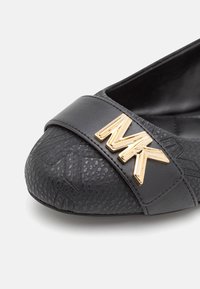 Chaussure plate noire texturée avec une large bride sur l'avant-pied, ornée d'un logo MK métallique doré.