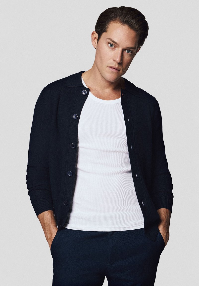 Granatowy sweter z dzianiny z dużymi guzikami, noszony na białym topie. Materiał jest teksturowany, a sweter ma kołnierz i dopasowane rękawy.