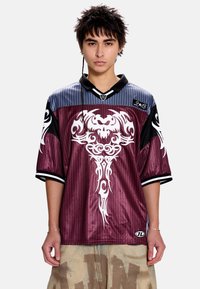 Maillot en maille bordeaux avec des accents noirs et bleus, arborant un motif tribal blanc complexe et un col classique. Manches courtes à rayures.