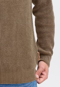 Brązowy sweter o teksturowanym wzorze, z dzianiny w kratkę, z prążkowanymi mankietami i dołem, z małą metką oznaczoną "INDICODE" na boku.