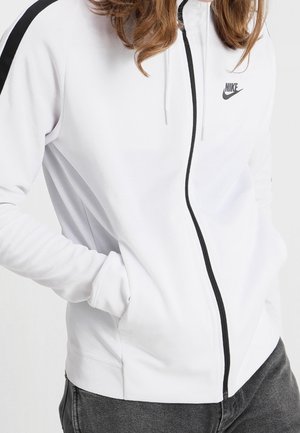 Personne portant une veste zippée blanche Nike avec une bande noire sur la manche et une fermeture éclair noire, les mains dans les poches, associée à un jean foncé.