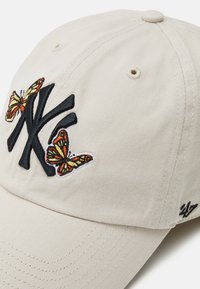 Beige baseballpet met zwart geborduurd "NY" logo en kleurrijke vlinderontwerpen. Gelaagd materiaal met een gebogen rand en een verstelbare band.