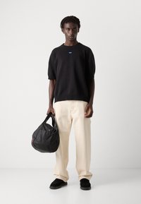 Pulover scurt, negru, cu maneci scurte, pantaloni crem, pantofi negri slip-on, ținând o geantă neagră cu un accent roșu, pe un fundal minimalist.