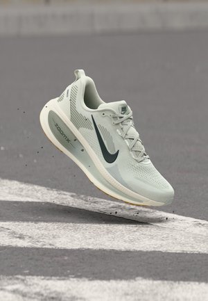 Lys grå Nike løpesko med svart swoosh, ZoomX-såle og lisser, svevende over en grå asfaltflate med hvite veimerkede striper.