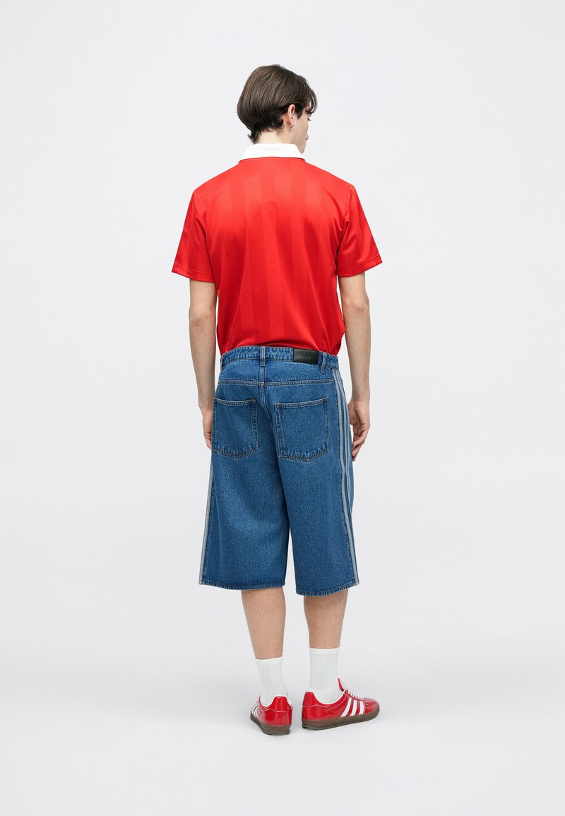 Jeune homme debout de dos, portant un polo rouge, un short en denim bleu jusqu'aux genoux, des chaussettes blanches et des baskets rouges avec des rayures blanches.