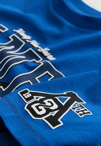 Blaues T-Shirt mit kurzen Ärmeln aus Baumwolle, mit weißer und schwarzer Schrift und einer Grafik in der unteren Ecke. Glatte Textur mit Rundhalsausschnitt.