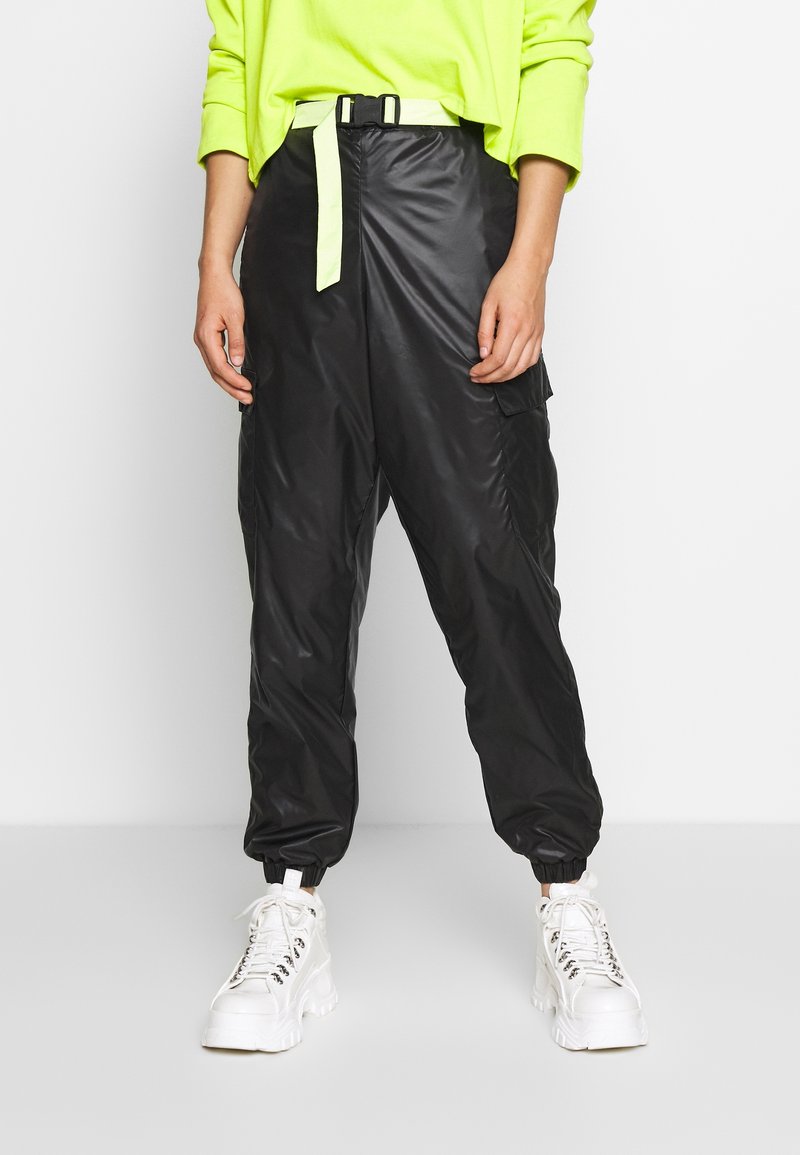 Svarta, glänsande, nylon cargo-byxor med elastiska muddar och ett lysande grönt bälte, kombinerat med kraftiga, vita sneakers.