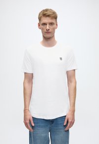 Bijela pamučna majica s okruglim izrezom i kratkim rukavima, s malim crnim logotipom na lijevoj strani prsa. Nošena s plavim trapericama.