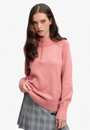 Vrouw met lang bruin haar, een roze trui met halve rits en een grijze geruite geplooide rok, staande met één hand die de zoom van de trui vasthoudt.