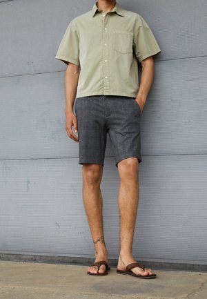 Korte mouw olijfgroen overhemd met knopen, grijze geruite korte broek en bruine slippers; model staat tegen een grijze achtergrond.