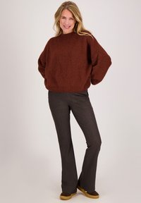 Lachende vrouw staat met handen achter de rug, draagt een dikke bruine trui, donkere uitlopende broek en bruine schoenen met rubberen zolen.