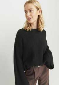 Suéter negro de punto acanalado con cuello alto y mangas anchas, combinado con pantalones marrones. La textura parece suave y cómoda.