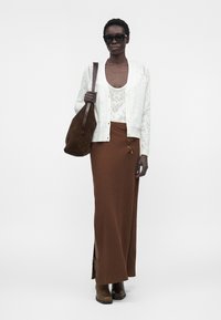 Femme portant des lunettes de soleil noires, un cardigan en dentelle blanche et un top, une longue jupe marron, des bottes marron, portant un grand sac à bandoulière en daim marron.