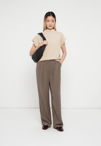 Top beige a maniche corte abbinato a pantaloni marroni a vita alta. Gli accessori includono una borsa nera a tracolla e scarpe bordeaux con lacci.