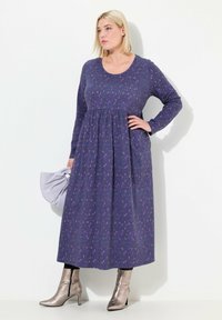Abito a maniche lunghe di colore viola con motivo floreale, vita arricchita e gonna lunga fino alla caviglia. La modella indossa stivaletti argentati e tiene una borsa grigia.