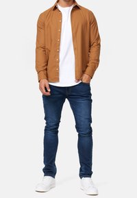 Braun langärmliges Hemd über einem weißen T-Shirt, kombiniert mit blauen Slim-Fit-Jeans und weißen Sneakers. Das Material wirkt glatt; Knöpfe sind sichtbar.