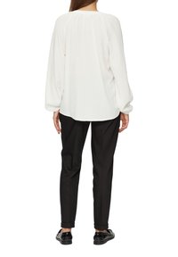 Blouse blanche texturée à manches longues, avec des détails froncés au col et une coupe décontractée, associée à un pantalon noir ajusté et des chaussures noires brillantes.