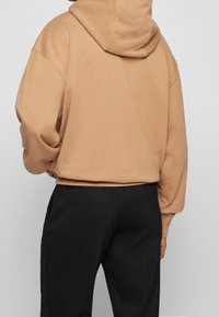 Persona con sudadera con capucha color beige combinada con pantalones negros, vista desde atrás sobre un fondo claro y liso.