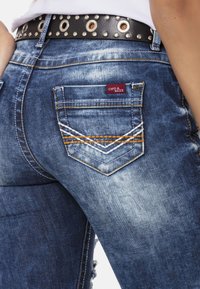 Denim spijkerbroek met een vervaagde blauwe wasbeurt, voorzien van een klein rood merklabel, drie keer gestikte achterzakken en een zwarte gesp met studs.