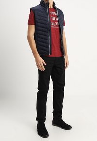 Marine mouwloze puffer vest over een rode graphic t-shirt, gecombineerd met zwarte slim-fit jeans en zwarte enkellaarsjes. Ritssluiting, gestikte textuur.