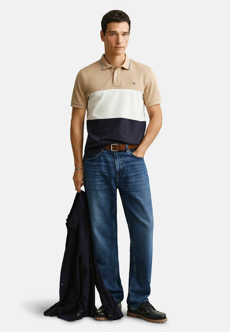 Homme portant un polo beige, blanc et bleu marine, jean bleu, chaussures noires, tenant une veste noire dans une main, debout devant un fond blanc.