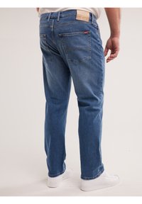 Vīrietis, kur š valkā zilas džinsa bikses un baltas sporta kurpes, redzams no aizmugures pret vienkāršu gaišu fonu.