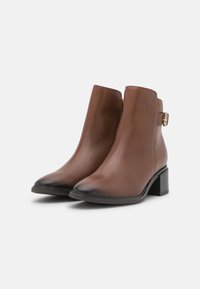 Botas de tobillo de cuero marrón con una textura suave, punta afilada, tacón ancho y cremallera lateral. Presenta una correa decorativa con detalle de hebilla.