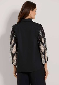 Schwarze Bluse mit durchsichtigen, bestickten Ballonärmeln, die ein Spitzenmuster aufweisen. Die Rückansicht zeigt einen strukturierten Kragen und einen glatten Stoff.