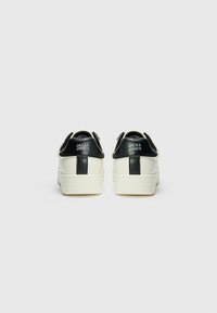 Witte sneakers met een gestructureerde afwerking, zwarte hielaccenten en logodetail. Ronde neus en platte rubberen zool met een profielpatroon.
