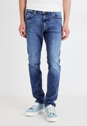 Man wearing slim-fit blue jeans and light blue sneakers standing against a plain white background.