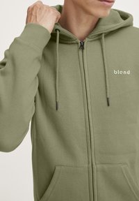 Felpa verde con zip realizzata in tessuto morbido, dotata di cappuccio regolabile con cordino, tasche frontali e un piccolo logo "blend" bianco sul petto.