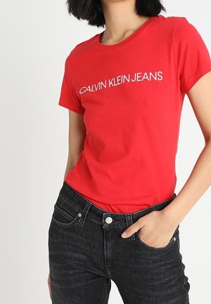 Czerwona bawełniana koszulka z okrągłym dekoltem i krótkimi rękawami, z białym napisem "CALVIN KLEIN JEANS". Założona z ciemnymi dżinsami.