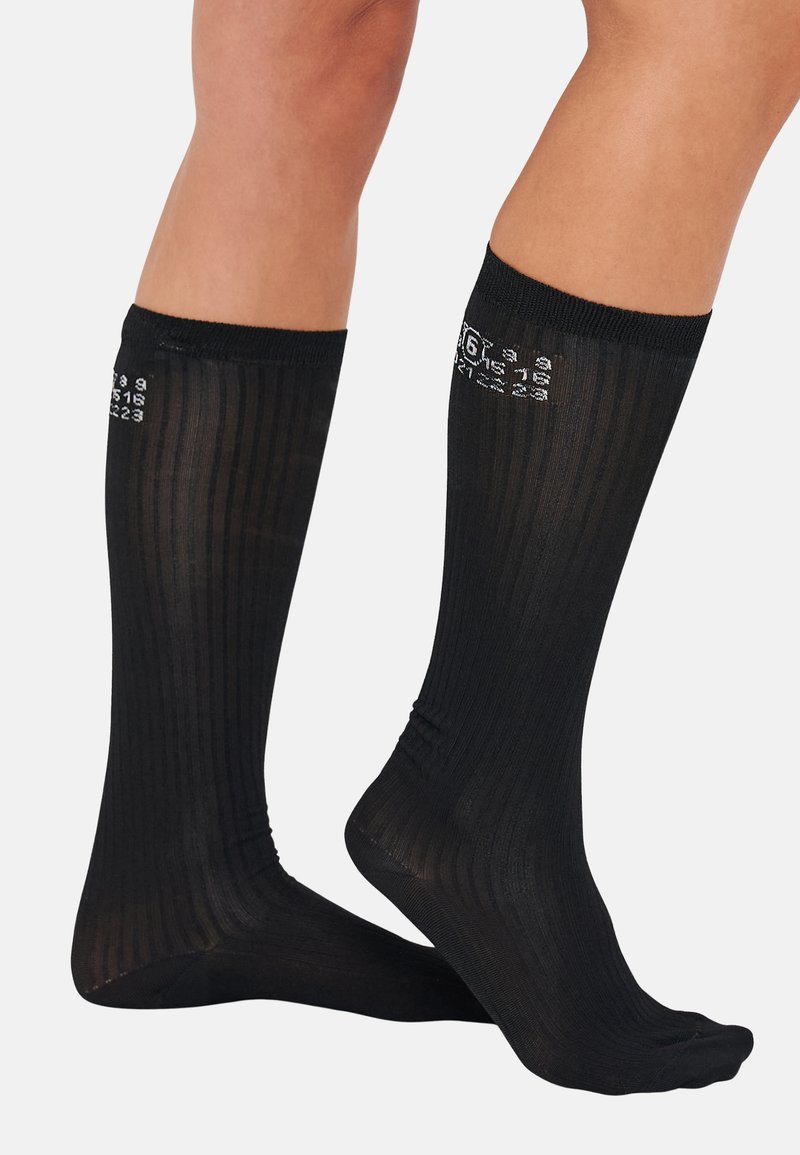 Chaussettes noires montantes en maille côtelée verticale, avec des chiffres et des lettres blancs imprimés sur le dessus. Design léger et semi-transparent.