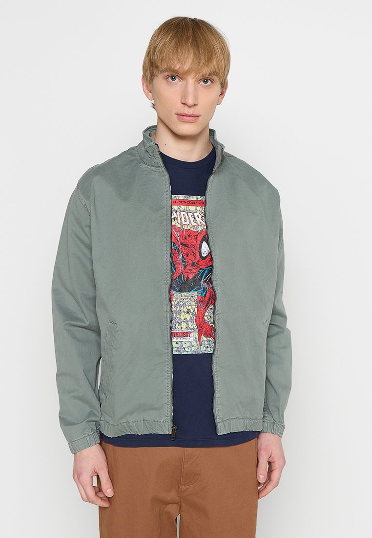 Hellegrüne Zip-Jacke mit Stehkragen, elastischen Bündchen und Saum, getragen über einem navyblauen T-Shirt mit Grafik in Comic-Design.