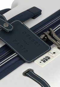 I bagagli Delsey Paris presentano una scocca rigida bianca, dettagli in pelle blu navy, un'etichetta per i bagagli e una combinazione di lucchetto con hardware argentato.
