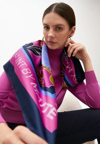 Farbenfroher Seidenschal, der über einem pinken Rollkragenpullover drapiert ist. Der Schal zeigt florale Muster und kontrastierende marineblaue Akzente mit Text.