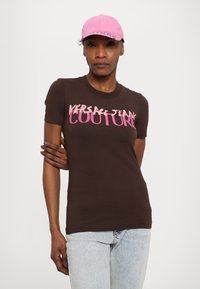 Brūns pieguļošs T-krekls ar gaiši rozā "VERSACE JEANS COUTURE" tekstu. Kombinēts ar gaiši rozā cepuri. Audums izskatās gluds, ar īsām piedurknēm.