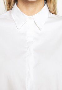 Blusa bianca con colletto, caratterizzata da una texture morbida, con chiusura a bottone e una forma classica. Il colletto è strutturato con punte appuntite e affilate.