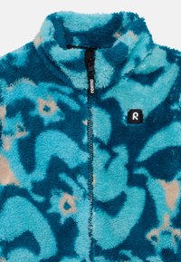 Chaqueta de forro polar en azul y verde azulado con patrones abstractos y un cuello alto. Presenta una cremallera negra con la marca "reima" y un parche de logo negro.