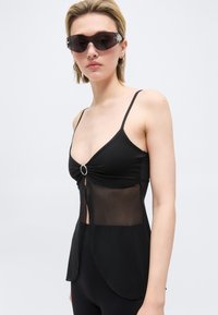 Γυναίκα με μαύρο αμάνικο τοπ με διάφανο τμήμα στη μέση και μεγάλα σκοτεινά γυαλιά ηλίου, μπροστά σε απλό ανοιχτό φόντο.