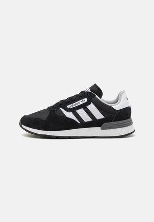 Black and gray sneaker made of mesh and suede, featuring three white stripes, lace fastening, and a textured rubber sole with branding on the heel.