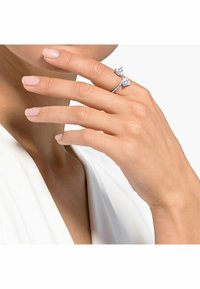 Zilveren ring van sterling zilver met twee hartvormige, heldere edelstenen, verfraaid met accentstenen langs de band, gedragen aan een hand met natuurlijke nagels.