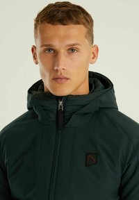 Temno zelena kapuca jakna z teksturirano površino, z zadrgo spredaj in logotipom na prsih. Visok ovratnik za dodatno toploto.