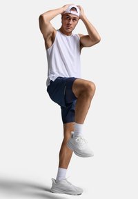 Biała koszulka bez rękawów, granatowe szorty z bocznymi siateczkowymi wstawkami, białe buty sportowe i biała czapka noszona do tyłu. Stojący na jednej nodze.