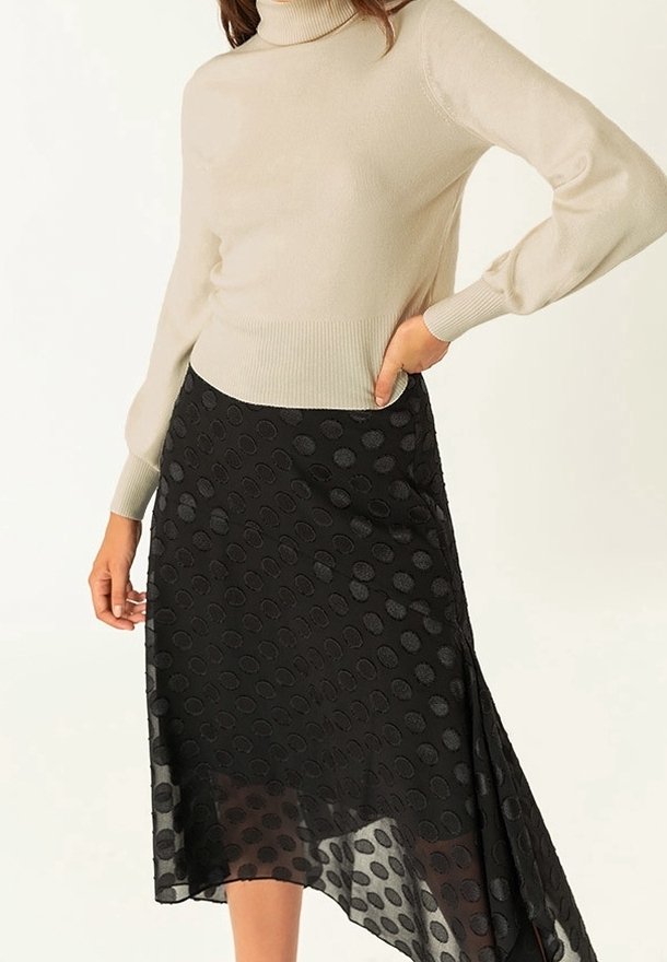 Beige Rollkragenpullover mit langen Ärmeln, kombiniert mit einem schwarzen asymmetrischen Rock, der ein strukturiertes Punktmuster aufweist.